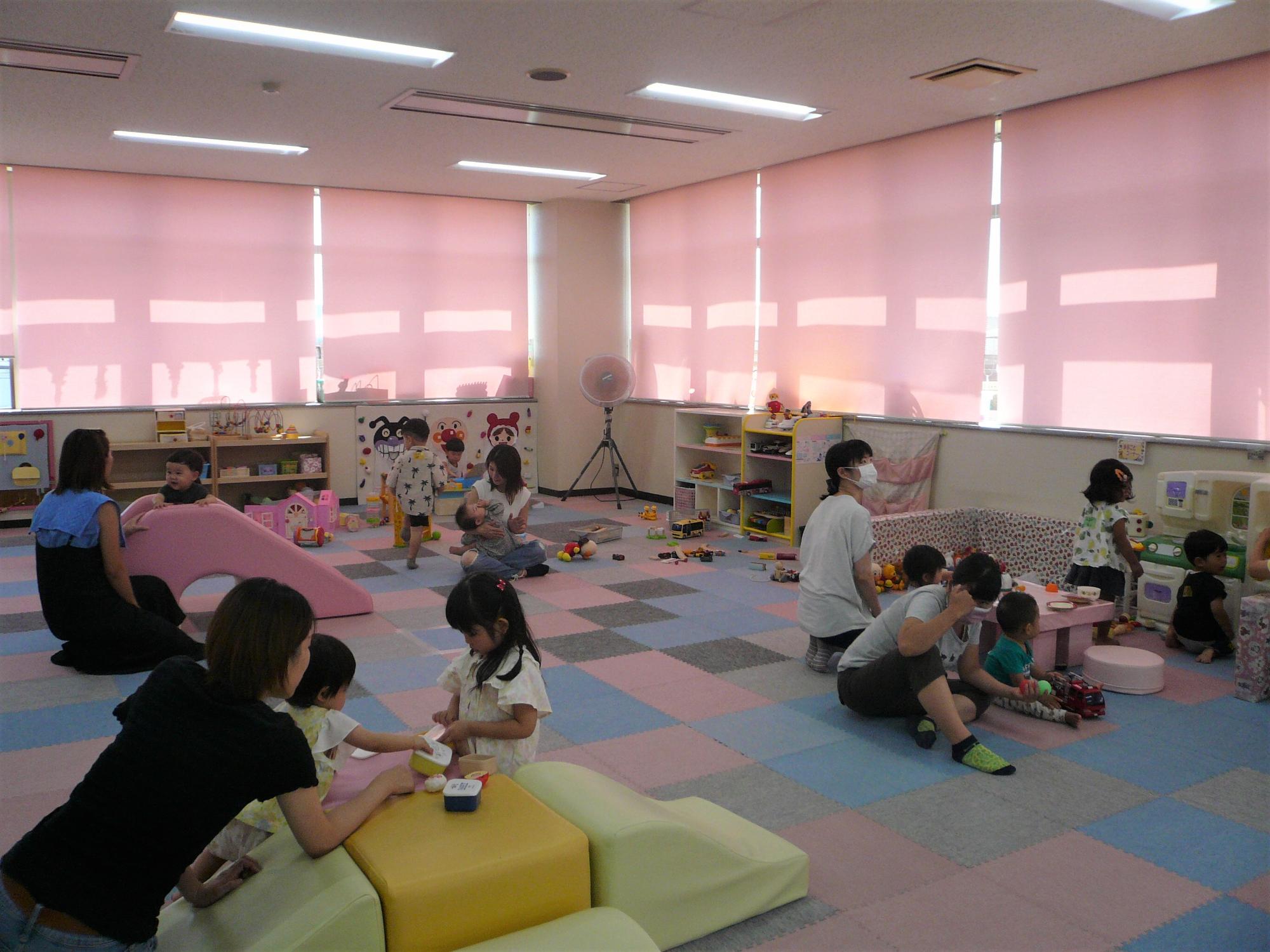 窓のある明るい室内の中、滑り台やブロックなど様々な遊具で遊ぶ子供達や、そばに座っている大人達の写真