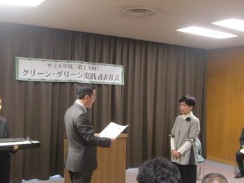 平成26年度クリーン・グリーン実践者表彰式にて、同表彰式の名前が書かれた横断幕の前で表彰状が読み上げられている様子の写真