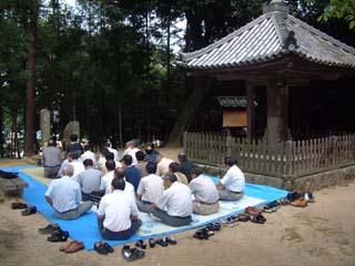 傘堂の脇にある2つの墓碑の前にブルーシートを広げて座り、法要を行う参列者たちの写真