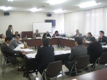左手前から撮った、四角く並べられた席を囲んで会議する人々の中で、角の人物がマイクを持ち立ちながら話している写真