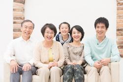 3世代の家族が前に大人達が座っていて後ろに女の子が顔を出して笑っている写真