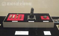 台の上に並べられた細長い棒状、直立する棒状、丸く平たい形の様ざまな縄文時代の祭祀具の展示風景の写真