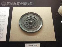 円形の皿の形で中央に丸い突起が並ぶ古い鏡が置かれた展示風景の写真