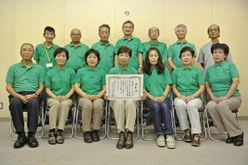 表彰状を持った肩を中央に二列に並んでいる、緑色の服を着た人々たちの集合写真