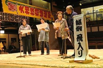 相撲館にて、マイクと甚句自慢飛入りと書かれためくりが用意された土俵の上に立っている人々の写真