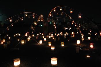 夜の暗闇のなか、たくさんのともしびが、据え置かれた地面やつりさげられた空中から、光を放っている写真