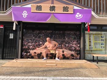 相撲館「けはや座」に設置された本場所の土俵と房を摸したフレームで土俵入りのポーズをまねている男性の写真