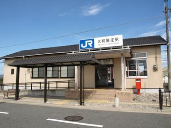 青空を背景に、赤いポストが置かれ、屋根にはJR大和新庄駅の駅名プレートが据えられている、白よりの壁に黒い屋根の駅舎の写真