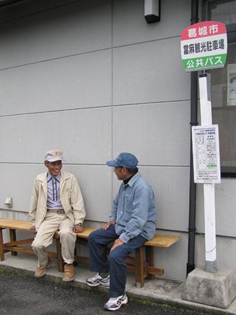 當麻観光駐車場のバス停の隣に設置された木製のベンチで男性2人が向かい合って話している写真