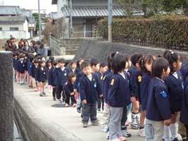 道路に並んで順番を待つ制服姿の低学年児童たちの列の写真