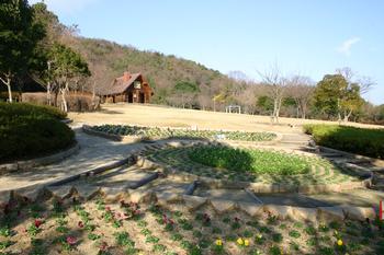 二上山ふるさと公園の花壇と芝生広場の写真。芝生公園の先にログハウス調のおもちゃ館が見える