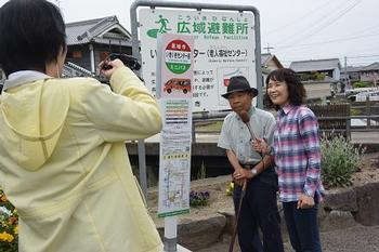 停留所の横でマイクを持った人と杖をついた人が立っていてそれを白いパーカーを着た人が撮影している写真