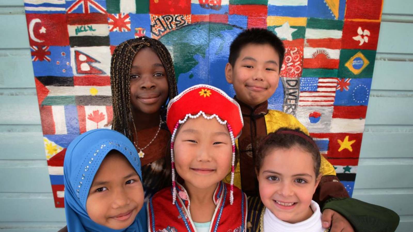 色々な国の国旗と世界地図が描かれている布の前で様々な衣装を着た4人の子供がいる写真