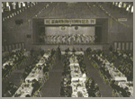 人々が立つステージが奥にあり、手前に縦に並べられたテーブルと、椅子に座る人々の白黒写真