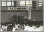 「アメニティ記念講演会」と書かれた幕が壁に貼られており、室内に人々が集まっている白黒の写真
