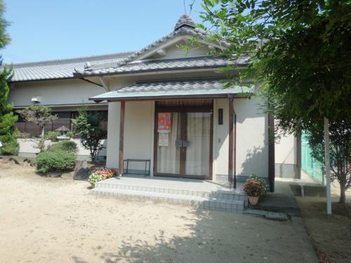 灰色の瓦屋根に白い壁の建物で花の植木が並べられている公民館今在家分館の入り口の写真